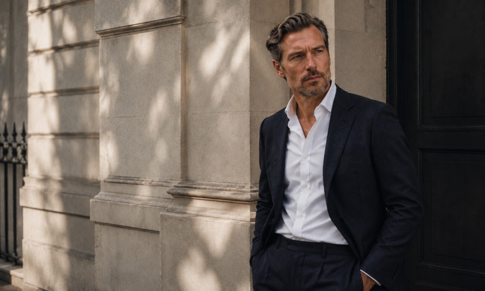 Man in a navy tailored jacket and white open-collar shirt leaning against a sunlit stone building, shop Richard James at Malford of London
