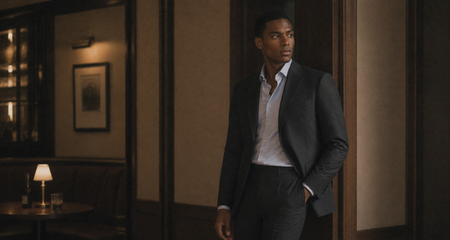 Man in a charcoal tailored suit and white open-collar shirt standing in a wood-panelled doorway, shop Tailoring at Malford of London
